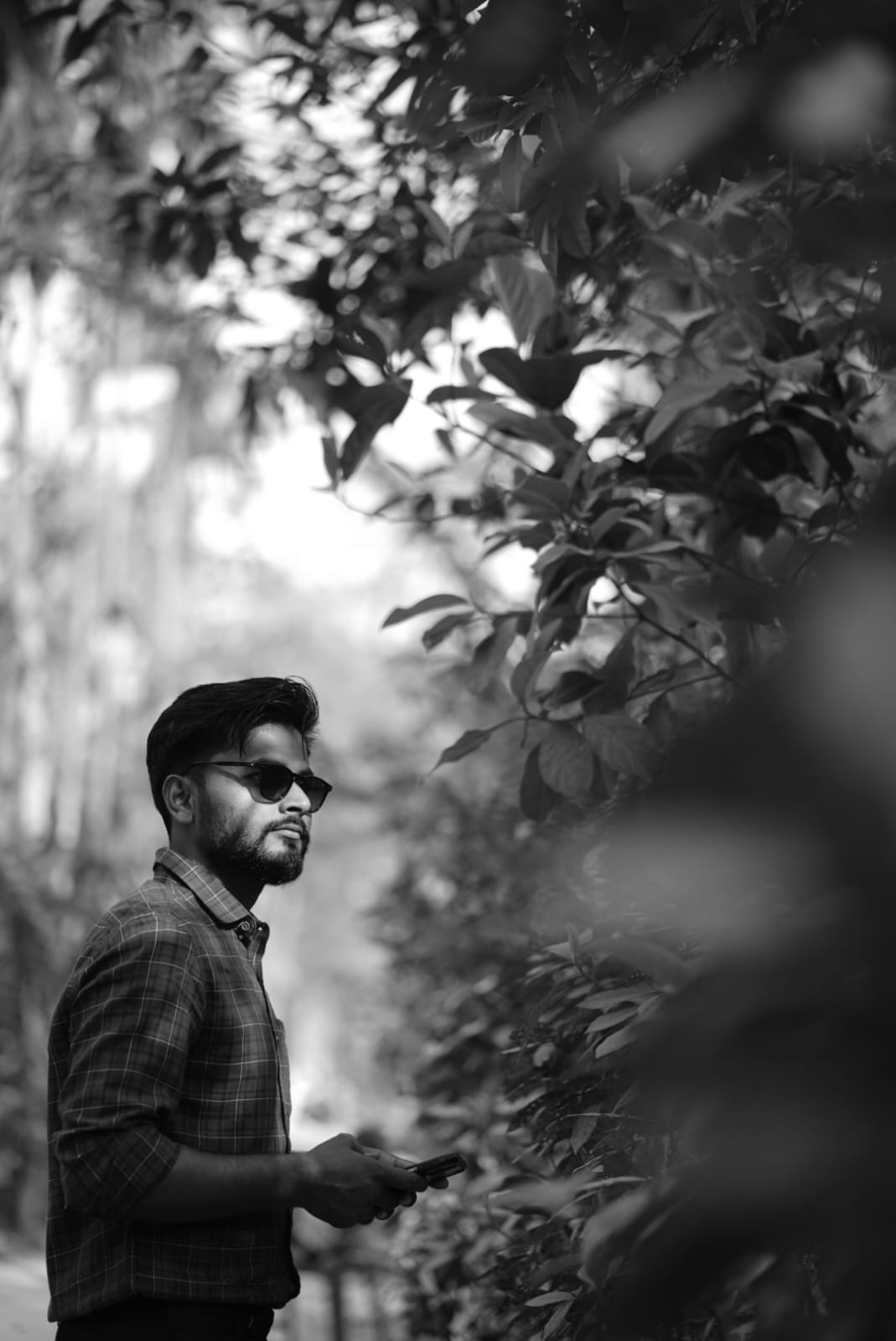 Rajesh wearing sunglasses stands in a lush green forest, surrounded by tall trees and dappled sunlight
